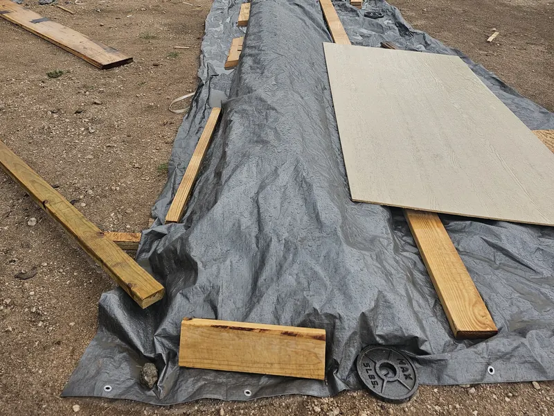 Photograph of lumber and board on the lot, covered by a tarp held down with weights.