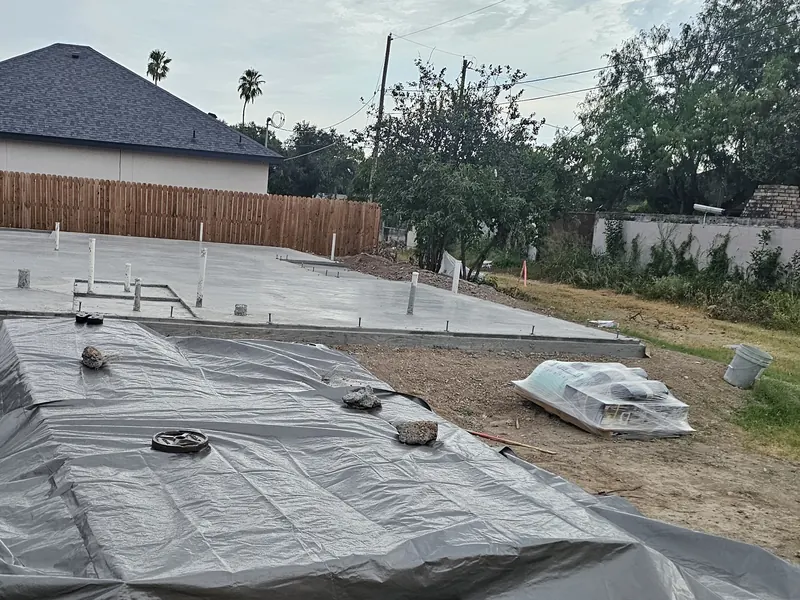Photograph of a freshly poured slab, with building materials under a weighted tarp in front of it.